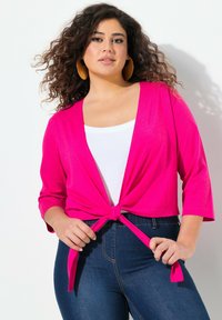 Cardigan court rose vif avec un détail à l'avant noué, des manches trois-quarts et une texture douce, superposé sur un haut blanc et un jean foncé.