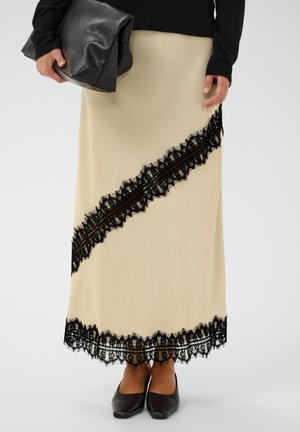 Jupe midi beige avec une bordure en dentelle noire diagonale et à l'ourlet, portée avec des chaussures pointues noires, un pull noir, et tenant une grande pochette noire.