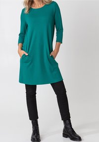 Teal, midi-jurk met een ronde halslijn en driekwartmouwen. Beschikt over zijzakken en gecombineerd met zwarte broek en veterschoenen.
