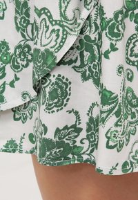 Vit tyg med intrikata gröna blommönster och paisleymönster, med en nederkant som har en subtil textur och kontrasterande grön kant.