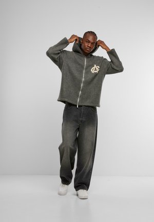 Grijze zip-up hoodie met een groot logo op de linkerkant van de borst, gecombineerd met losse zwarte denim broek en witte sneakers.