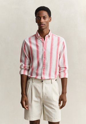 Mann trägt ein weißes und rosa gestreiftes Hemd mit aufgekrempelten Ärmeln und beigefarbene plissierte Shorts, steht vor einfarbigem Hintergrund.
