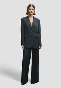 Donker krijtstreeppak met een op maat gemaakte blazer met een opstaande revers en bijpassende broek met brede pijpen. Beide stukken zijn gemaakt van een gestructureerde stof.