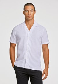 Camisa blanca de lino de manga corta con cuello de solapa, con un frente abotonado y tejido texturizado. Combinada con pantalones negros.