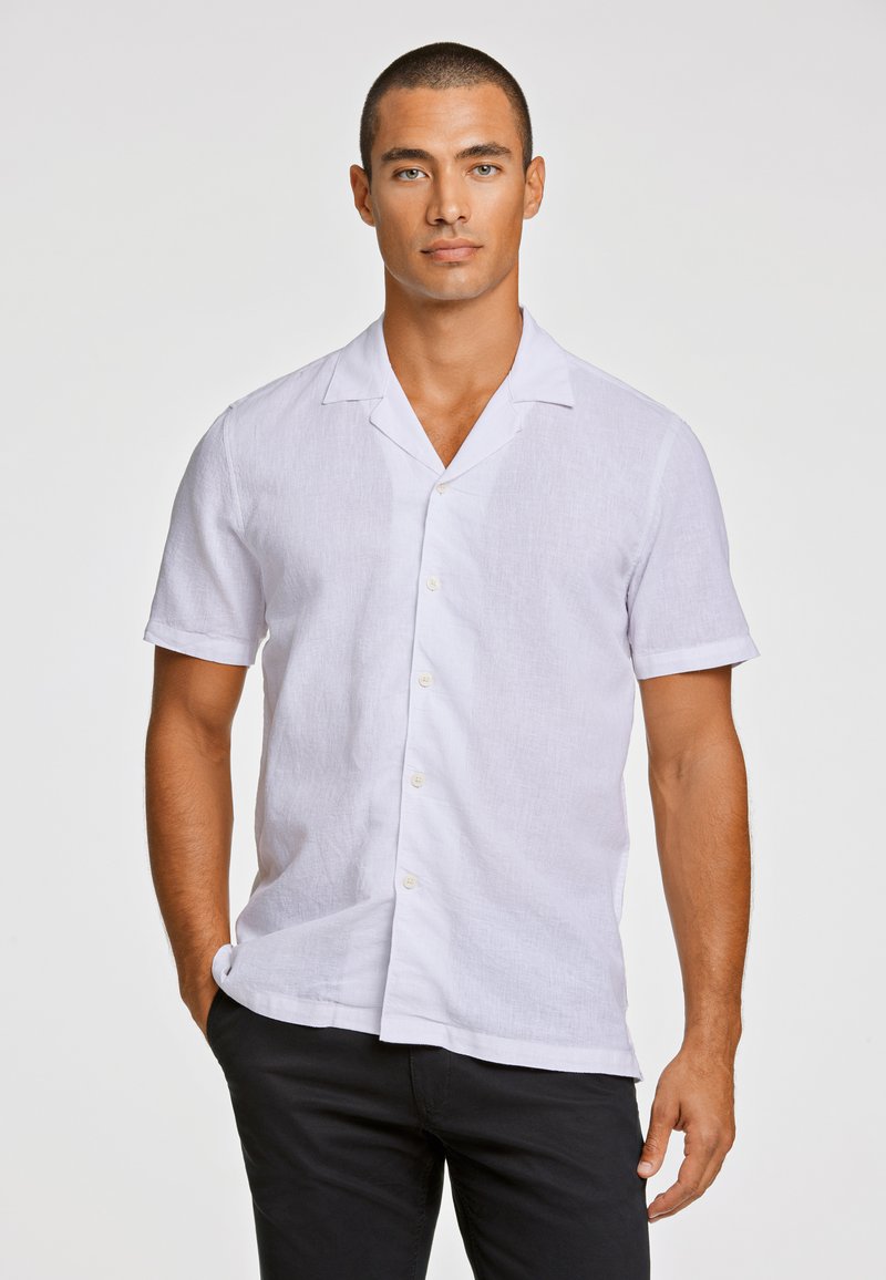 Camisa blanca de lino de manga corta con cuello de solapa, con un frente abotonado y tejido texturizado. Combinada con pantalones negros.