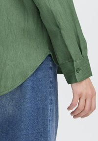Main reposant à côté d'un jean en denim bleu, portant une chemise verte à manches longues avec un poignet boutonné et un ourlet légèrement incurvé.