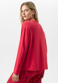 Vrouw met blond haar kijkt over haar schouder, gekleed in een rode blouse met lange mouwen en rode broek met prints van zwarte cartoonfiguren.