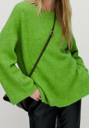 Kobieta ubrana w jasnозielony, oversize, puszysty sweter z szerokimi rękawami, czarne skórzane spodnie oraz czarny pasek torby przewieszony przez klatkę piersiową.