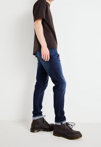 T-shirt corto a maniche corte di colore marrone scuro, jeans blu slim-fit e stivaletti in pelle marrone a caviglia con lacci neri e cuciture gialle su uno sfondo chiaro.