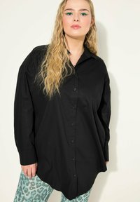 Sort oversized skjorte med knaplukning foran, lange ærmer, blødt stof og buet kant, parret med lyse teal leopardmønstrede leggings.
