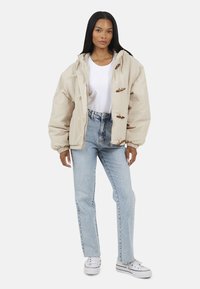 Beżowa, oversize'owa kurtka z kapturem, z zapięciem na toggle, noszona na białej koszuli i jasnoniebieskich jeansach w kształcie dzwonów.