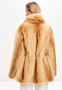 Faux fur frakke i lys tan med en fyldig tekstur, der har en høj krave og en flagrende silhuet. Sømdetaljer fremhæver formen.