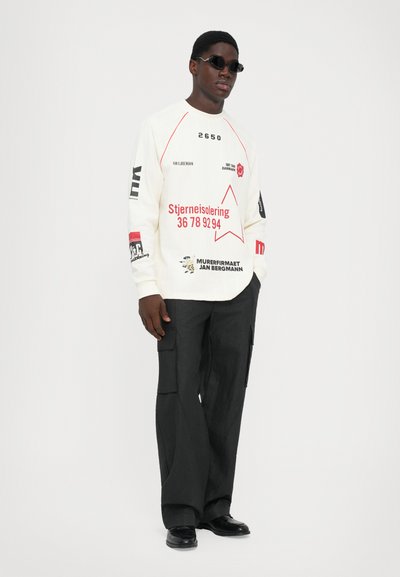 Man wearing white long-sleeve shirt with red and black text, black cargo pants, black shoes, and small black sunglasses on white background.