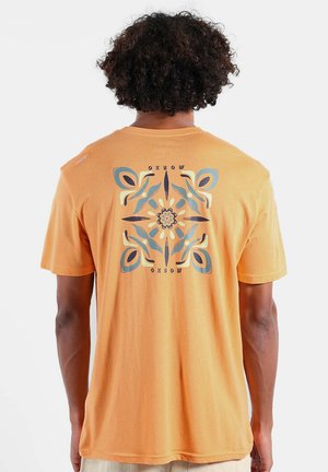 Personne aux cheveux bouclés portant un t-shirt orange avec un motif floral abstrait bleu et crème et le texte "OXBOW" au dos.