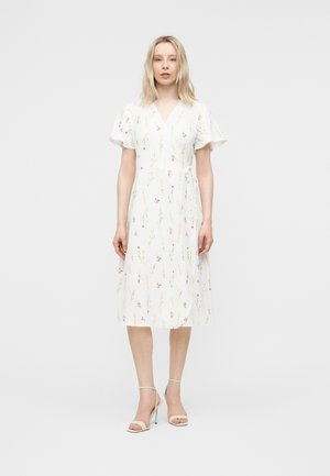 Kvinde iført hvid midi slå-om kjole med blomsterprint og flagrende ærmer, stående mod en ensfarvet lys baggrund med et neutralt udtryk.