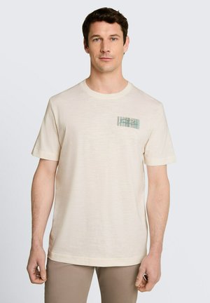 Mężczyzna noszący jasnobeżowy t-shirt z krótkim rękawem z małą prostokątną zielono-beżową paskowaną naszywką na lewej piersi.