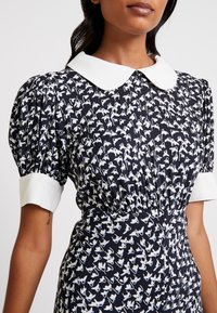 Robe bleu marine avec manches bouffantes et col blanc, présentant un motif de petits oiseaux bleu clair et blancs. Taille froncée pour une silhouette ajustée.