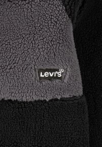 Črna in siva flis tkanina z teksturirano površino. Črni logo "Levi's" na sivem delu. Brez dodatnih okrasitev.
