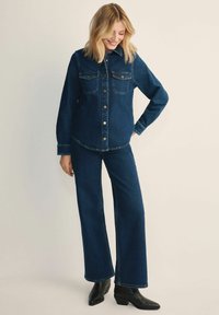 Zestaw skórzanej kurtki i spodni dzwonów w ciemnym niebieskim kolorze. Kurtka posiada dwie kieszenie na klatce piersiowej, zapięcie na guziki oraz klasyczny kołnierz. Botki w czarnym skórzanym wykończeniu.