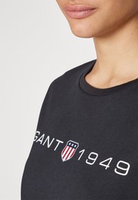 Nærbillede af person iført sort crew neck t-shirt med teksten "GANT 1949" og skjoldlogo med stjerner og striber.