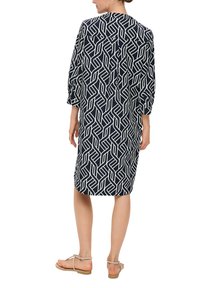 Marineblaues Kleid mit einem weißen geometrischen Muster, Dreiviertelärmel, knielang, aus leichtem Material und mit seitlichen Schlitzen, kombiniert mit Sandalen.