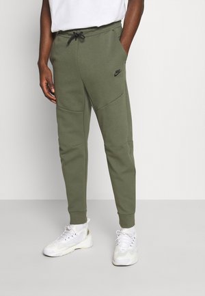 Mann trägt olivgrüne Nike Jogginghose mit Kordelzugbund, weißes Shirt, weiße Socken und weiße Sportschuhe und steht mit den Händen in den Taschen.