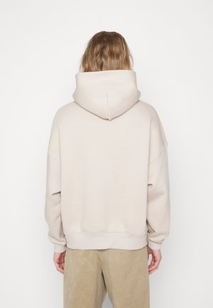 Osoba z długimi włosami, ubrana w beżową bluzę z kapturem i jasnobrązowe spodnie, stojąca przodem do białego tła, z rękami swobodnie opuszczonymi po bokach.