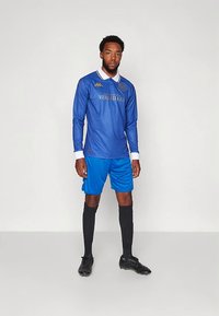 Blå långärmad fotbollströja med vit krage, med gyllene bokstäver och logotyp. Bärs med blå shorts, svarta strumpor och svarta fotbollsskor.