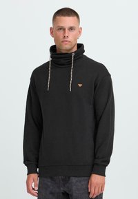 Hombre con cabello corto lleva un sudadera negra de cuello alto con cordones, de pie contra un fondo claro y liso, mirando hacia adelante con expresión neutral.