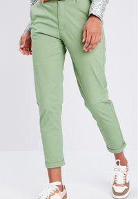 Pantalon vert clair à coupe droite, ourlets retroussés et texture lisse. Associé à des baskets blanches et marron.