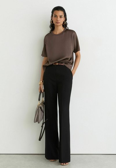 Woman wearing brown short-sleeve top, black high-waisted wide-leg pants, holding grey handbag, standing against white wall indoors.