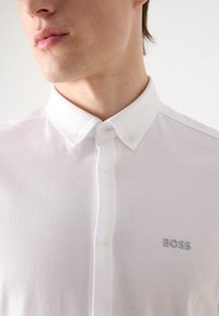 Camicetta bianca con colletto button-down, maniche corte e una sottile trama. Presenta un piccolo logo 'BOSS' grigio chiaro sul petto.