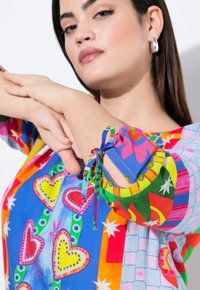 Blouse colorée présentant des motifs vibrants avec des cœurs et des étoiles. Base bleue, accents multicolores, et manches à nouer ajustables avec des découpes.