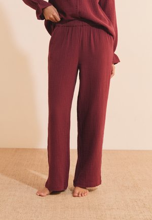 Personne portant un pantalon ample, texturé, rouge foncé, debout pieds nus sur un sol recouvert de moquette claire, une main ajustant la taille.