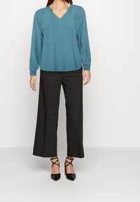 Blouse teal à manches longues avec un col en V et des poignets élastiques, associée à un pantalon à jambes larges noir et des escarpins noirs à bout pointu avec des brides à la cheville.