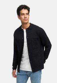 Ung mand iført en sort knappet cardigan over en hvid T-shirt og blå jeans, der ser let til siden med en neutral udtryk.
