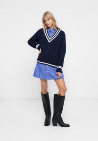 Navy gestrickter Pullover mit weißer Borte, über einem blau-weiß gestreiften Hemdkleid getragen, kombiniert mit knielangen schwarzen Stiefeln mit Absatz.