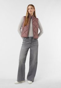 Gepolsterte ärmellose Weste in Staubrose, mit hohem Kragen und zwei seitlichen Reißverschlusstaschen. Kombiniert mit hellgrauen, weit geschnittenen Jeans und weißen Sneakers.