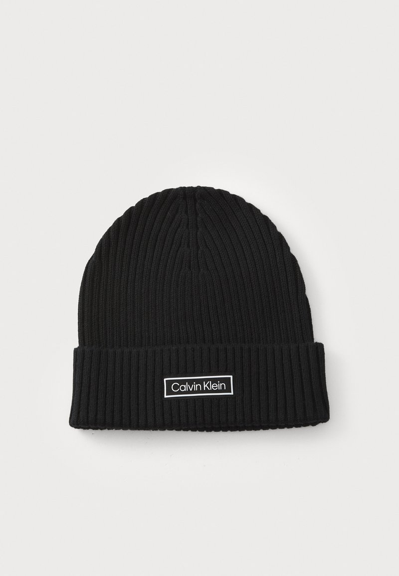 Sort strikket beanie med ribbet tekstur og opsmøg. Har et hvidt rektangulært mærke med "Calvin Klein" logo.