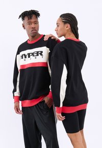 Čierna mikina s červenými a bielymi pruhmi, s logom "HYPER BOOST". Mäkká textura s rebrovanými manžetami a lemom. Nosené čierne športové šortky.