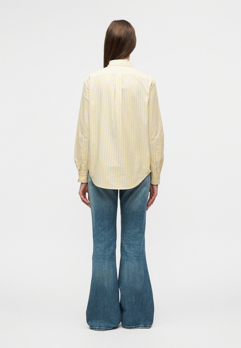 Chemise à rayures jaunes avec un col et des manches longues, associée à un jean bleu évasé. La chemise présente un ourlet arrondi et une coupe décontractée.