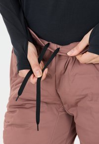 Roze gekleurde broek met een trekkoordsluiting, die door een hand wordt aangepast. De stof lijkt glad met een lichte textuur.