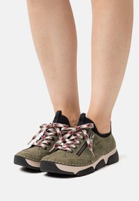 Baskets en suède vert avec accents noirs, dotées de semelles texturées, de lacets tissés colorés et d'un détail de fermeture éclair sur le côté.