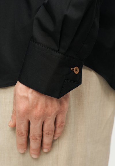 Ανδρικό χέρι που ακουμπάει δίπλα σε μπεζ παντελόνι, φορώντας μαύρο μανίκι πουκαμίσου με καφέ μανσετίνα με κουμπιά.