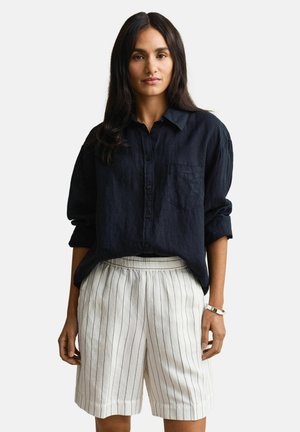 Vrouw met lang donker haar, gekleed in een donkerblauw overhemd met knopen en witte knielange shorts met dunne verticale strepen, staand met ontspannen armen.