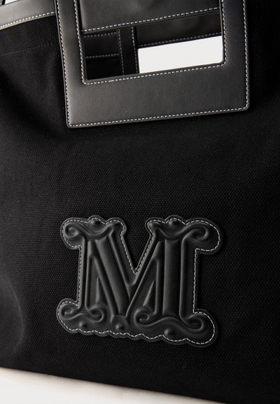 Μαύρη υφασμάτινη τσάντα με ορθογώνια δερμάτινη λαβή και μεγάλο ανάγλυφο δερμάτινο γράμμα "Μ" ραμμένο μπροστά.