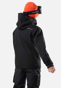 Svart vattentät jacka med huva och justerbara ärmmuddar, parat med svarta byxor. Har en logotyp på ryggen och en orange mössa. Snöskyddsglasögon ingår.