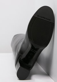 Botte en cuir noir montante montrant une semelle texturée avec logo. Bout rond et léger talon, finition lisse avec un détail de couture verticale.