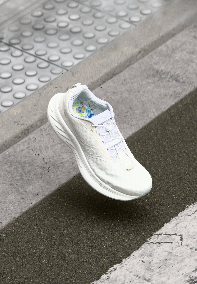Weiße Lauf sneaker mit strukturiertem Obermaterial, Schnürverschluss und blumigem Innensohle. Gebogene Sohlenform, in der Luft über Asphalt gezeigt.