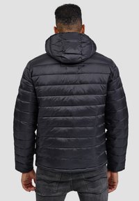 Chaqueta puffer negra con paneles acolchados horizontales, capucha y textura suave y brillante. Combinada con jeans gris oscuro.